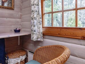 A cabin interior with a wooden wall, a window with a view of trees, a wicker chair, a small table with a blue cloth, and a curtain with a floral pattern. - 6 Person Holiday Home in Vaeggerlose (Vaeggerlose)