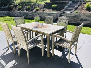 Una mesa de madera al aire libre con cuatro sillas, rodeada por un muro de piedra y una zona cubierta de césped.