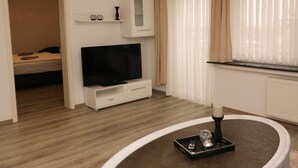 Un salon moderne avec un téléviseur à écran plat, un petit meuble, une fenêtre avec des rideaux légers et une table basse ronde sur laquelle se trouvent une télécommande et une bougie.