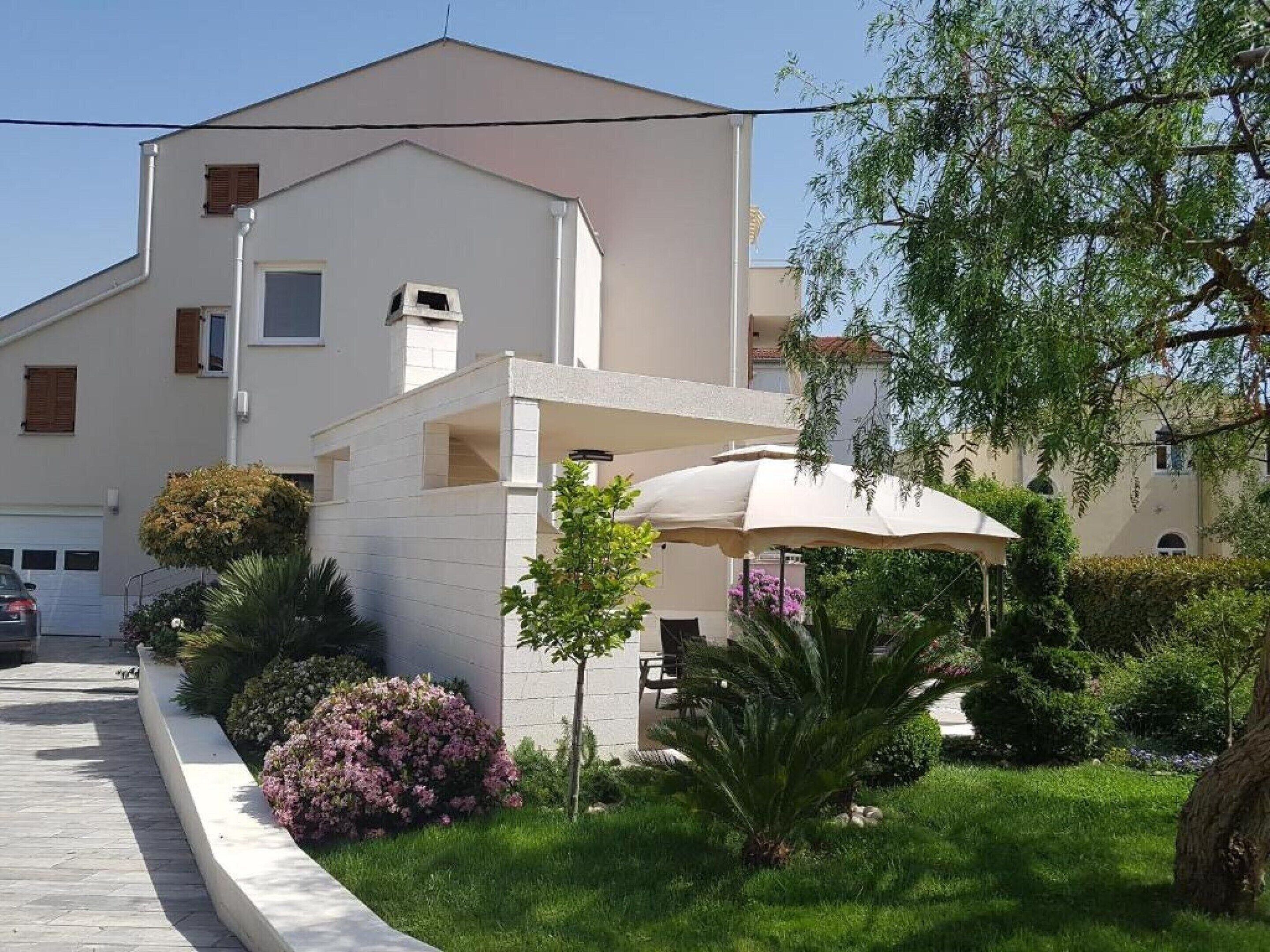 A modern two-story building with a flat roof, a covered outdoor seating area, and a well-maintained garden with various plants and a palm tree.