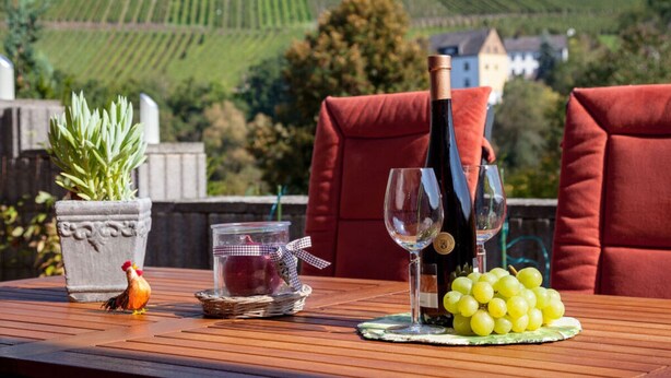A table set outdoors with a bottle of wine, two wine glasses, a bunch of grapes, a small jar with a candle, and a rooster figurine. - Holiday Home Moselle Valley (Maring-Noviand)