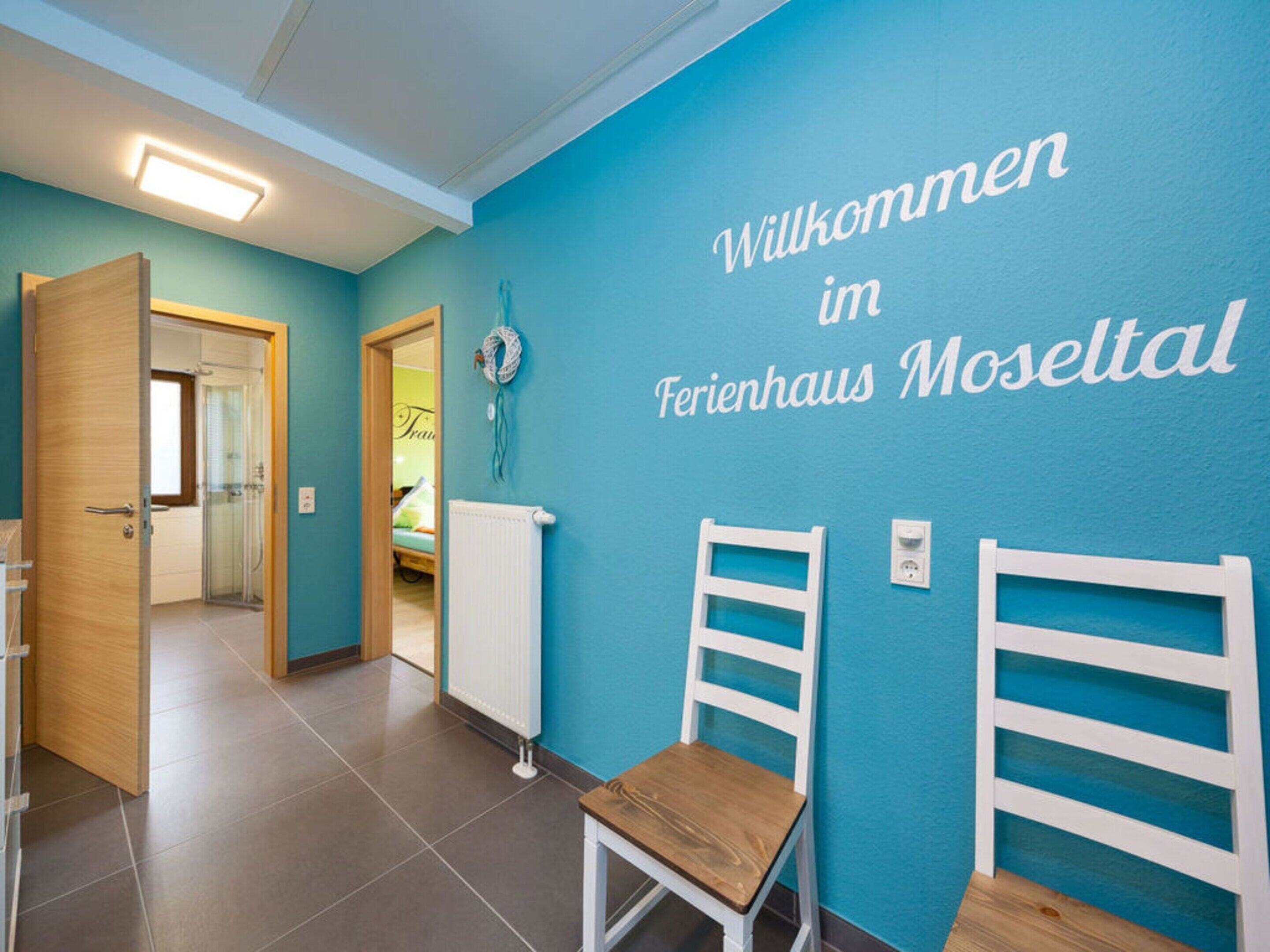 A hallway with a blue wall and a sign that reads "Willkommen im Ferienhaus Moseltal". There are two white chairs and a wooden chair in the hallway.