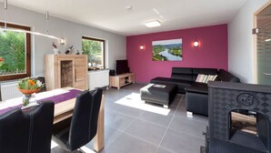 A modern living room with a black sofa, a dining area with a wooden table and chairs, and a fireplace. - Holiday Home Moselle Valley (Maring-Noviand)