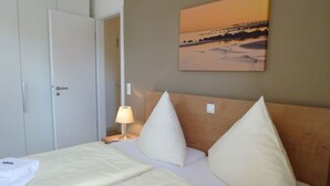 A hotel room with a bed, a nightstand with a lamp, a door, and a painting on the wall. - Deck 4 Modern Retreat (Timmendorfer Strand)