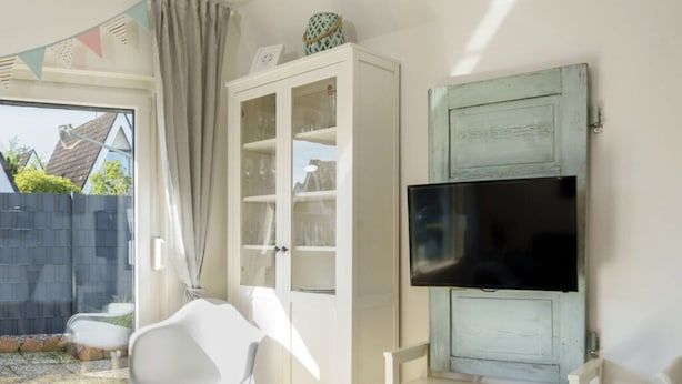 A room with a white cabinet, a TV mounted on a wall, and a white chair. - Holiday Home Heimathafen Am Ringwall (Cuxhaven)