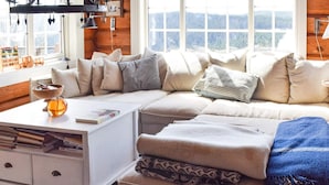 A cozy living room with a sectional sofa, a white side table, and a view of a forest through large windows. - 6 Person Holiday Home in Hurdal (Hurdal)