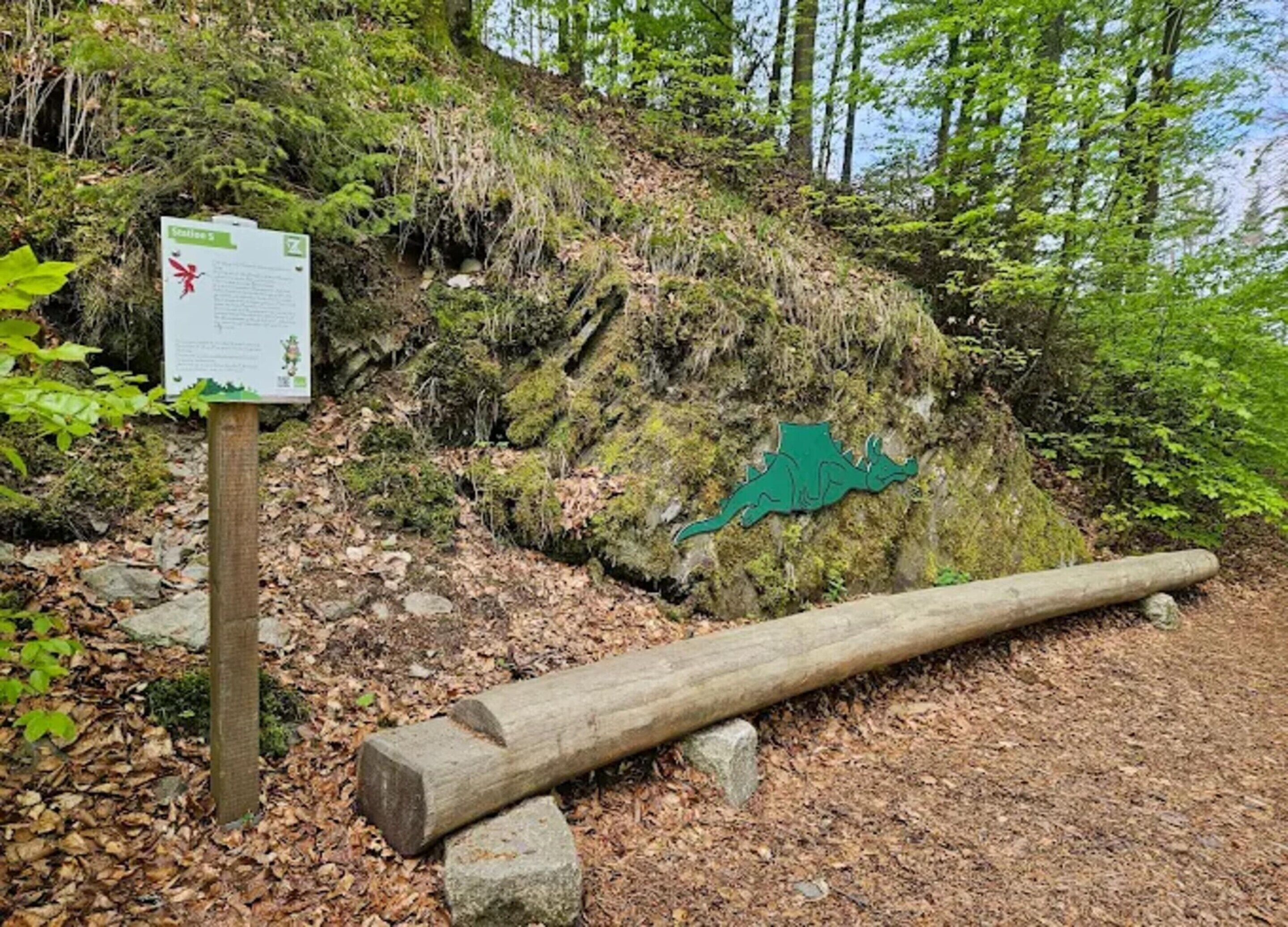 A natural setting with a wooden signpost, a large rock with a green dinosaur carving, and a fallen log.
