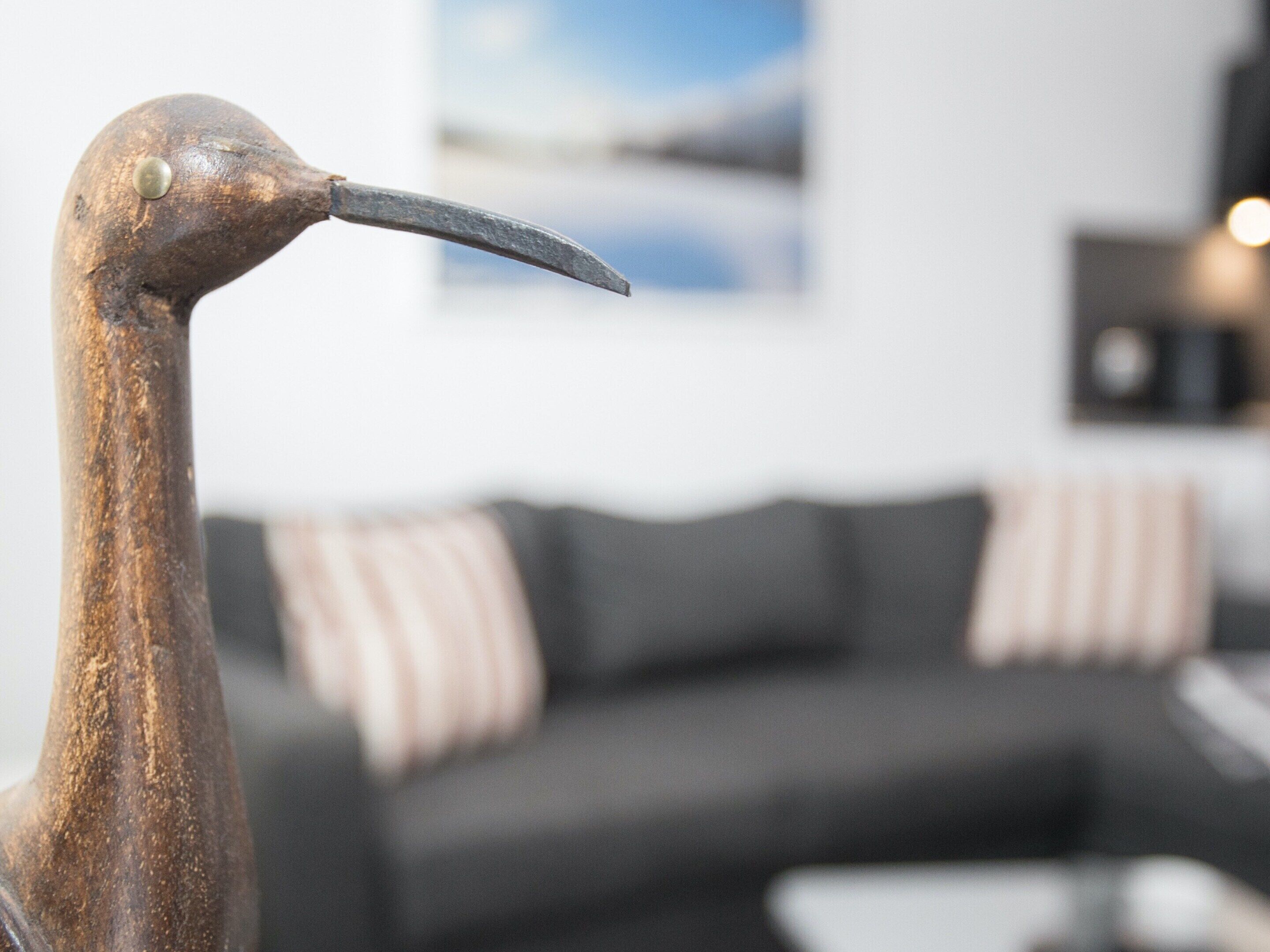 A close-up of a wooden sculpture with a curved beak, set against a blurred background featuring a dark-colored sofa with striped cushions.