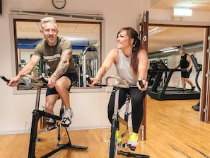 Duas pessoas pedalando em bicicletas ergométricas numa academia, com equipamentos de ginástica ao fundo.