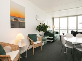 A modern living room with a sofa, chairs, a dining table, a lamp, a clock, and a large window with a view.