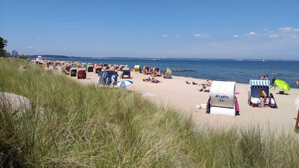 Une plage dotée de nombreuses chaises longues et de parasols, de quelques cabanes de plage, et offrant une vue dégagée sur la mer.