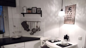 Une petite cuisine avec des meubles blancs, un plan de travail noir et une table ronde avec des chaises. On trouve au-dessus du plan de travail un porte-ustensiles suspendu et, au mur, un tableau d’acuité visuelle encadré.