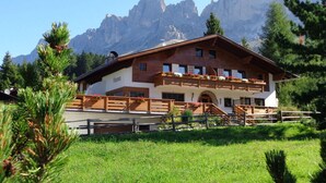 Un chalet à deux étages doté d’un balcon, entouré d’une clôture en bois et de verdure, avec une chaîne de montagnes en arrière-plan.