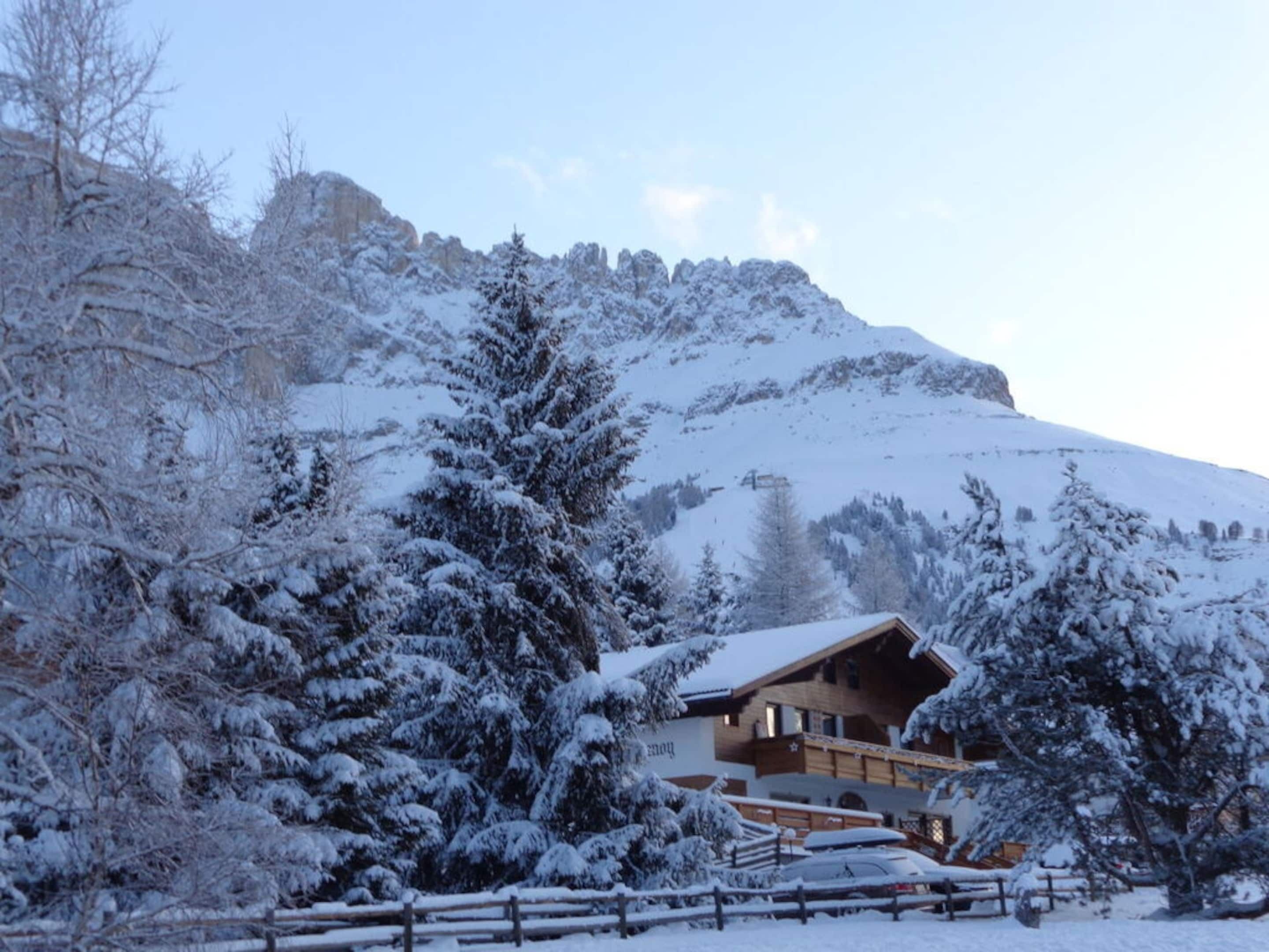 A wooden chalet with a sloped roof, surrounded by snow-covered trees and a mountain backdrop.
