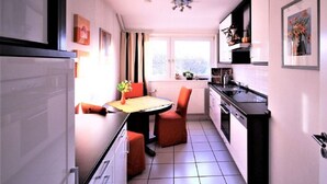 A compact kitchen with a dining table and chairs, a refrigerator, and a window with curtains. - Holiday Apartment 2 Oldenbug (Oldenburg)