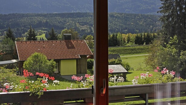 Une vue d’une maison avec un toit rouge et une porte verte, entourée d’un jardin où fleurissent des fleurs roses et blanches, et, au loin, une chaîne de montagnes boisées.