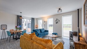 A modern living room with a sofa, a coffee table, and a fireplace. There is a dining area with a table and chairs, and a large window providing a view of the outdoors. - Holiday Home Near Forest With Wellness Garden (Den Burg)