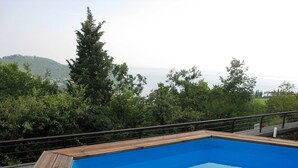 A rectangular pool with a wooden deck and metal railing, surrounded by lush greenery and a view of distant hills. - Holiday Home in Marciaga With Pool & Gulf Views (Costermano sul Garda)