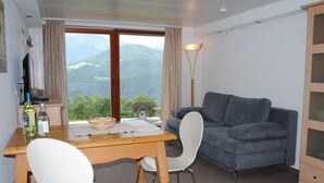 Une pièce avec une table en bois, deux chaises blanches, un canapé gris et une vue sur les montagnes, visible à travers de grandes fenêtres.