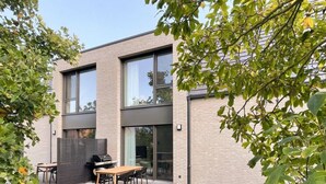Ein modernes, zweistöckiges Haus mit einer Terrasse, ausgestattet mit Tisch, Stühlen, einem Grill und einer Glastür zu einem überdachten Außenbereich.