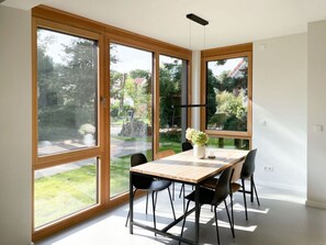 Un coin repas avec une table et des chaises en bois, de grandes fenêtres et une vue sur un jardin et des arbres.