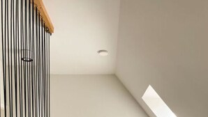 Ein modernes, minimalistisches Zimmer mit schrägen Wänden, einer Holzbank, einem Beistelltisch mit Büchern und einem Glas sowie einer Stehlampe.