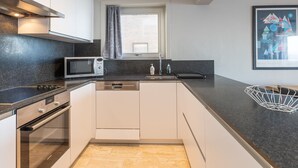 Una cocina moderna con microondas, horno y lavavajillas integrados. Hay una ventana con cortinas, un fregadero y un cuadro en la pared.