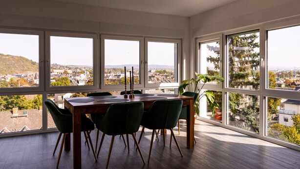 A dining area with a wooden table and chairs, large windows with a view of a cityscape and hills. - Apartment for 6 Modern Retreat (Andernach)