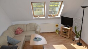 A modern living room with a beige sofa, a coffee table, a TV on a wooden stand, and two skylights with a view of greenery. - Holiday Apartment Nature View (Feldberger Seenlandschaft)