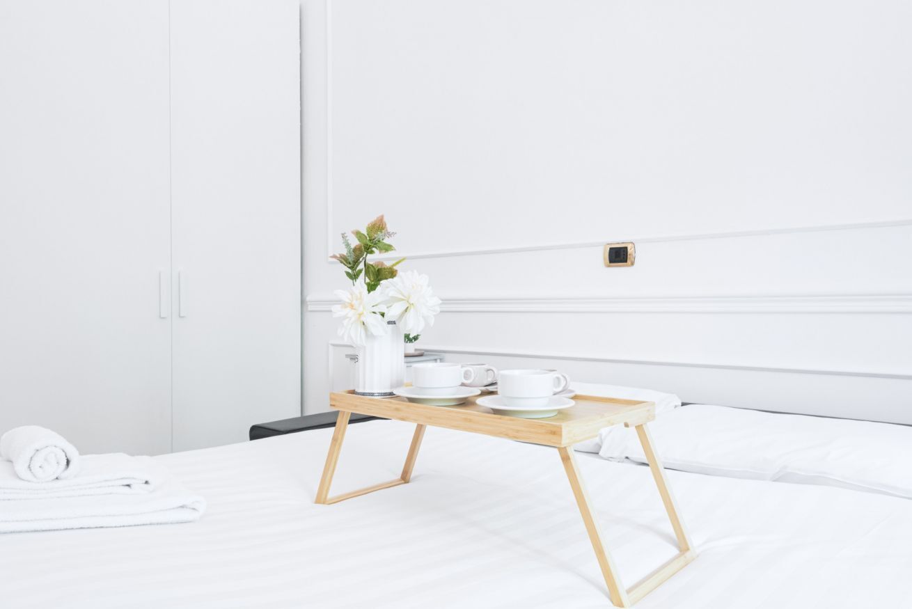 Un lit bien fait, avec une table de chevet en bois sur laquelle se trouvent un vase blanc rempli de fleurs, une bouilloire blanche et deux tasses blanches.