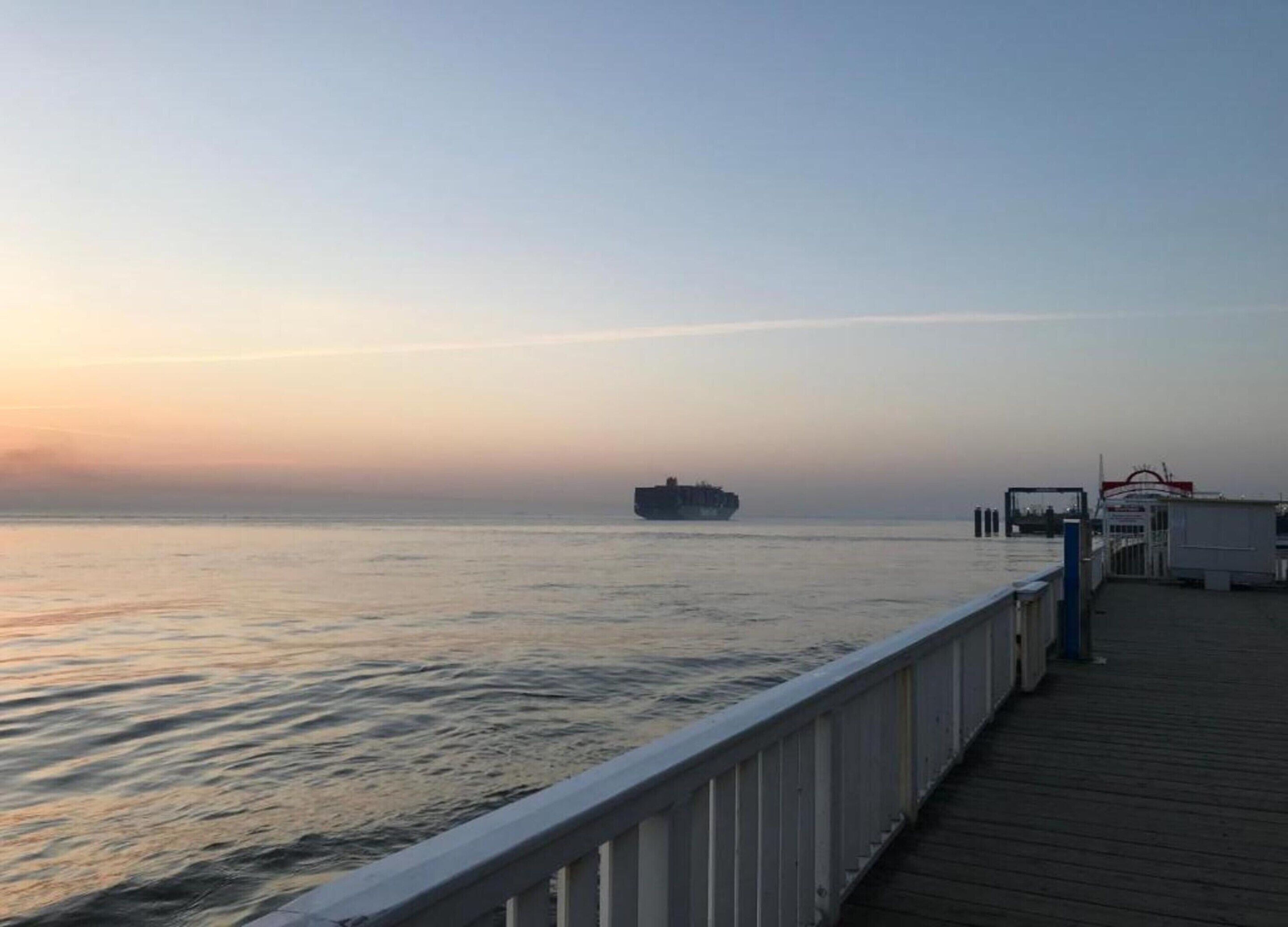 Une jetée avec un navire visible au loin, un bateau amarré et un petit bâtiment avec une enseigne rouge.