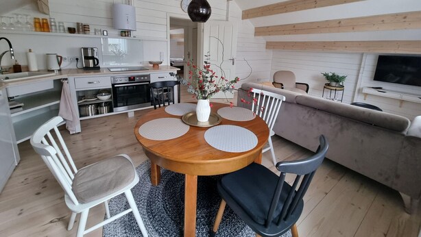 A modern kitchen with white cabinets, a built-in oven, and a coffee maker. There is a dining area with a wooden table and chairs, and a gray sofa in the living room. - Comfort Steps From the North Sea (Cuxhaven)