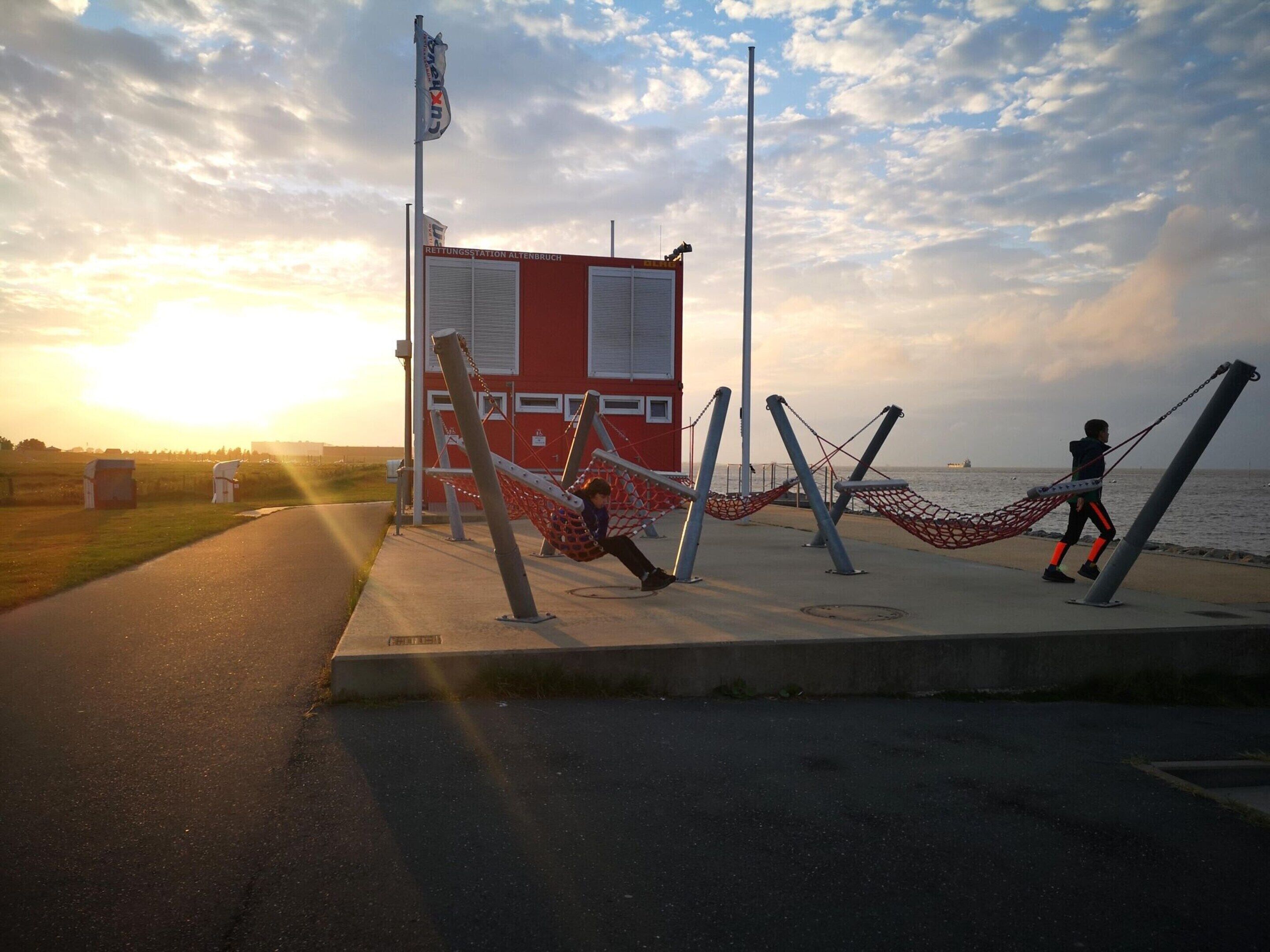 A red building with a sign reading "RETTUNGSTATION ALTENBRUCH" near a body of water. There are swings and a hammock on a concrete platform.