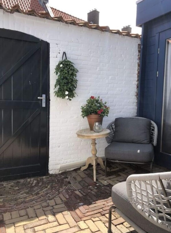 Un espace patio avec un mur en briques blanches, un portail noir, un pot de fleurs, une petite table ronde et une chaise grise.