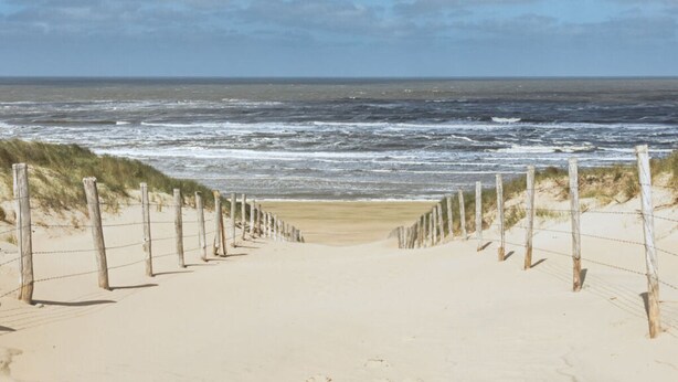 Un sentier de sable qui longe la plage et mène à l’océan, bordé de poteaux en bois et de fil de fer barbelé.