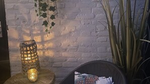 Une chaise en osier avec un coussin, une lanterne posée sur une table en bois et une plante suspendue devant un mur de briques.