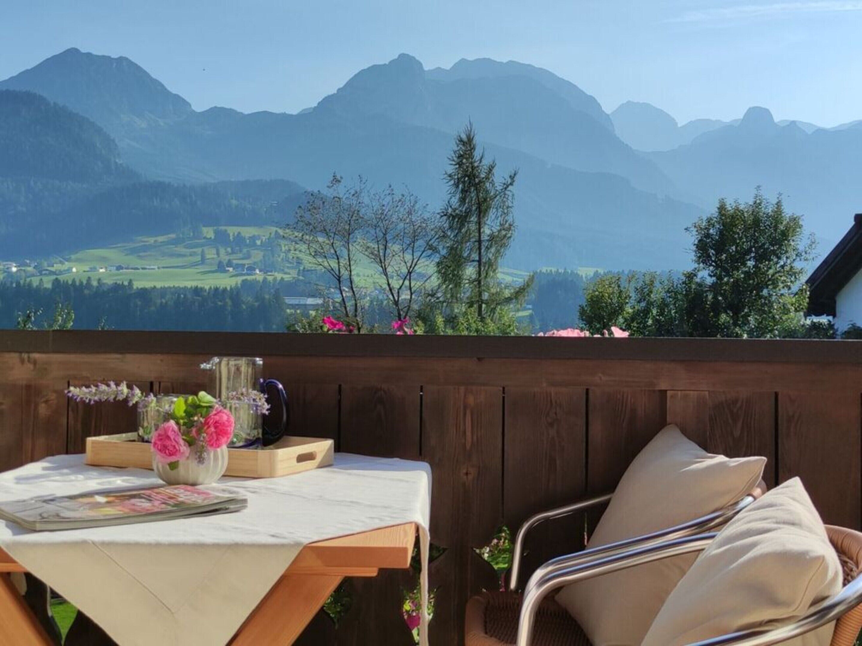 A wooden deck with a table set for two, a chair, and a vase of flowers. A mountain view is visible in the background.