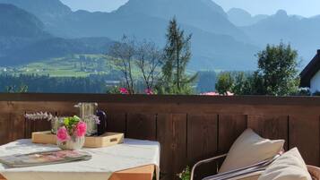 A wooden deck with a table set for two, a chair, and a vase of flowers. A mountain view is visible in the background.