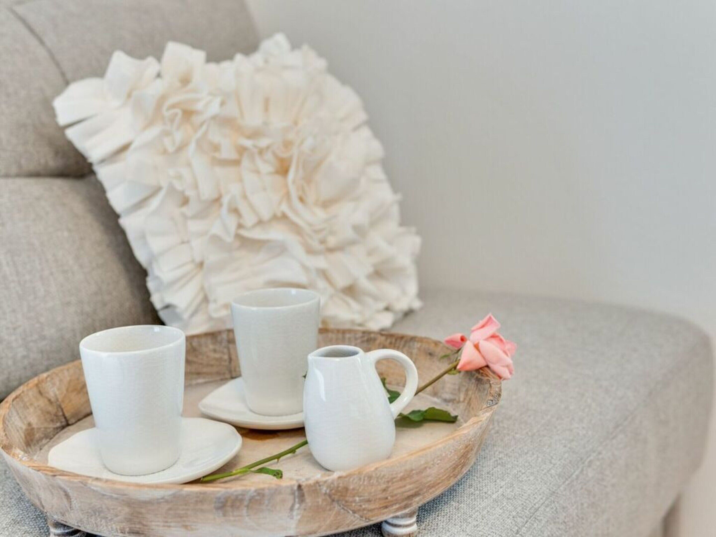 A tray with white cups and a pitcher on a wooden surface, placed on a grey sofa with a decorative pillow in the background.