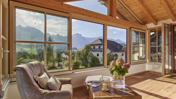 A spacious room with large windows offering a mountain view, a leather sofa, and a wooden coffee table. - Holiday Apartment The Neuhaus (Abtenau)