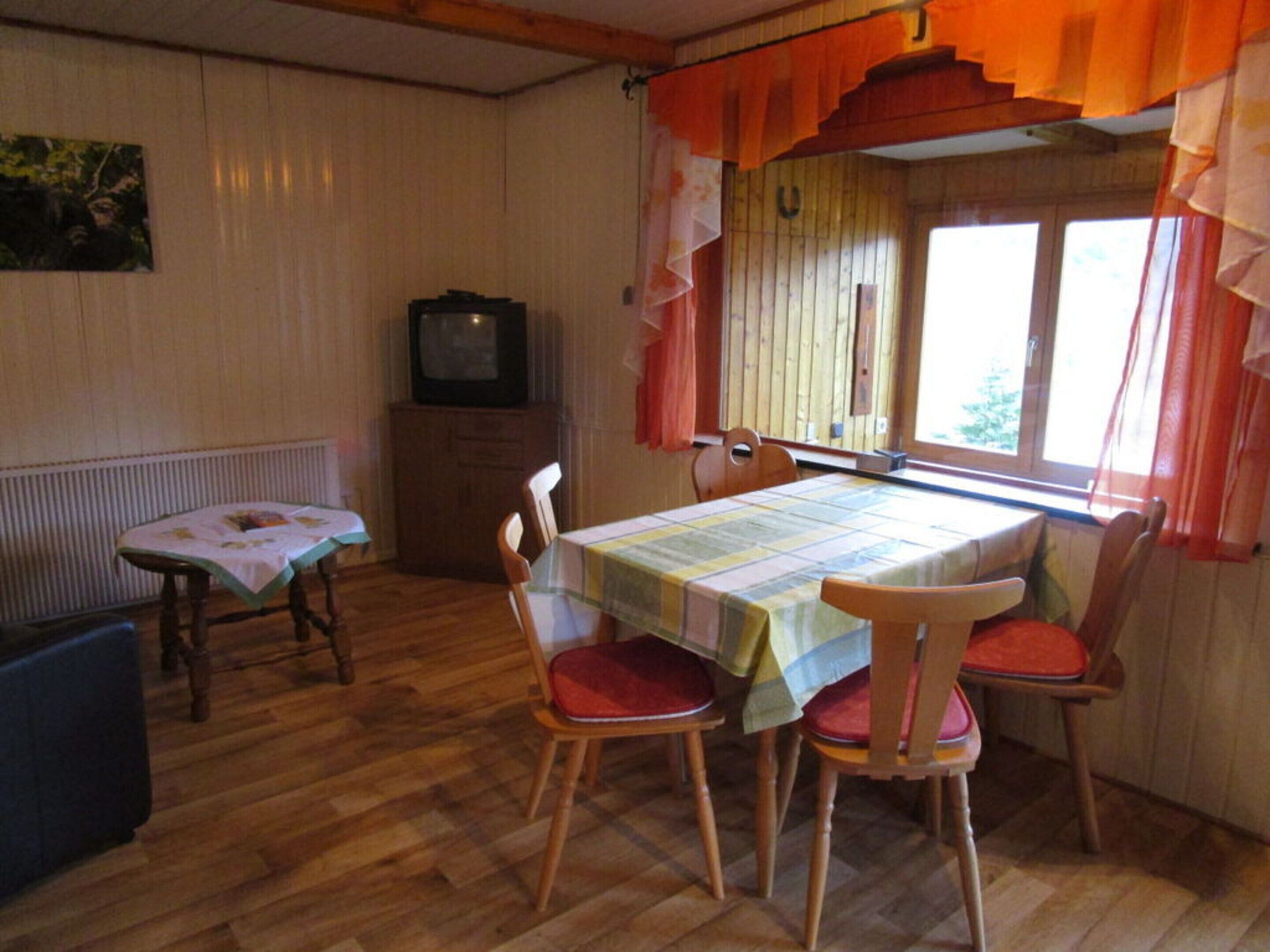 Een eethoek met een tafel voor vier personen, houten stoelen, een radiator, een televisie en een raam met gordijnen.