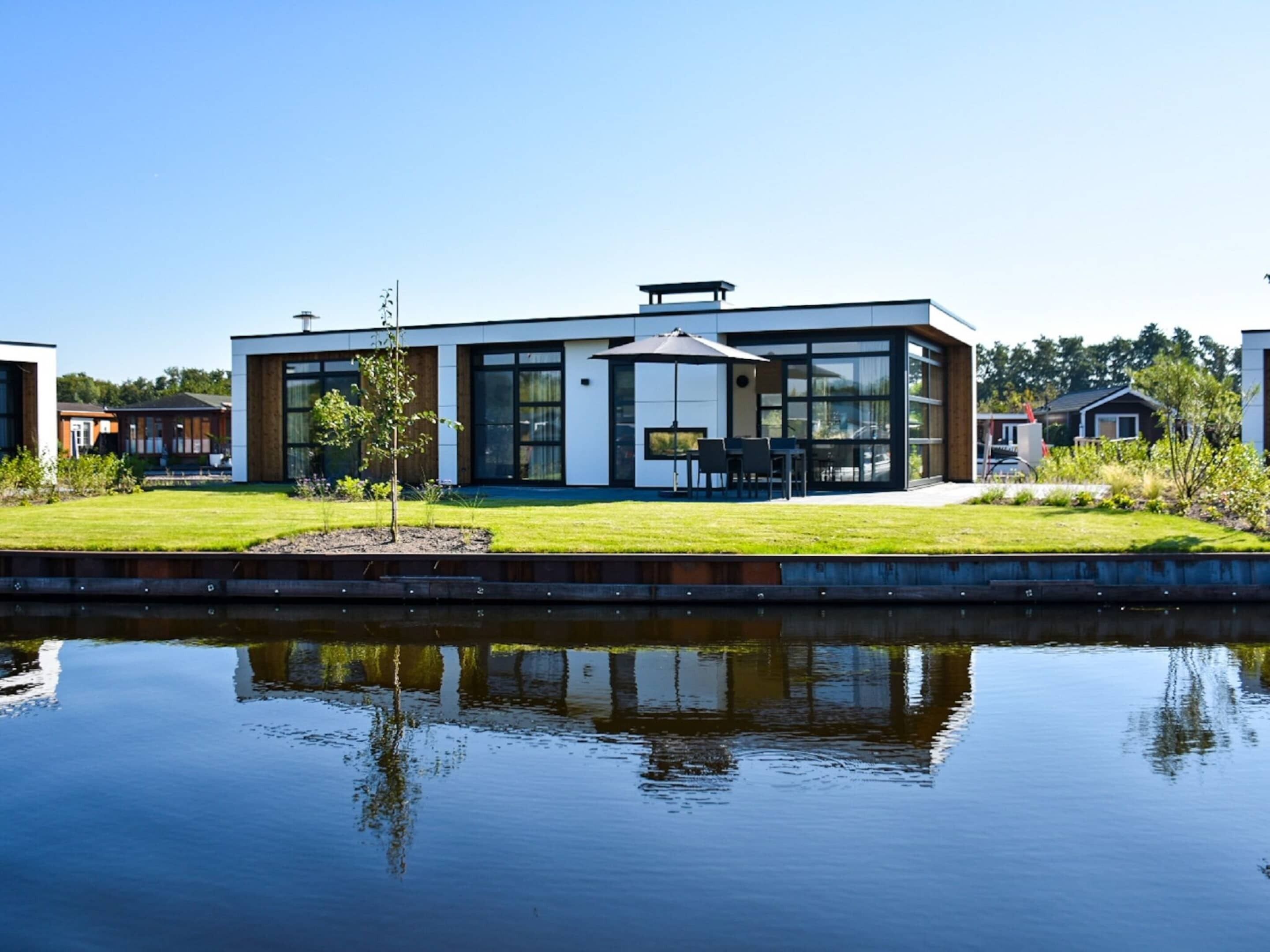 Une maison moderne à deux étages, dotée de grandes fenêtres, d’un patio avec mobilier d’extérieur et d’un petit jardin.