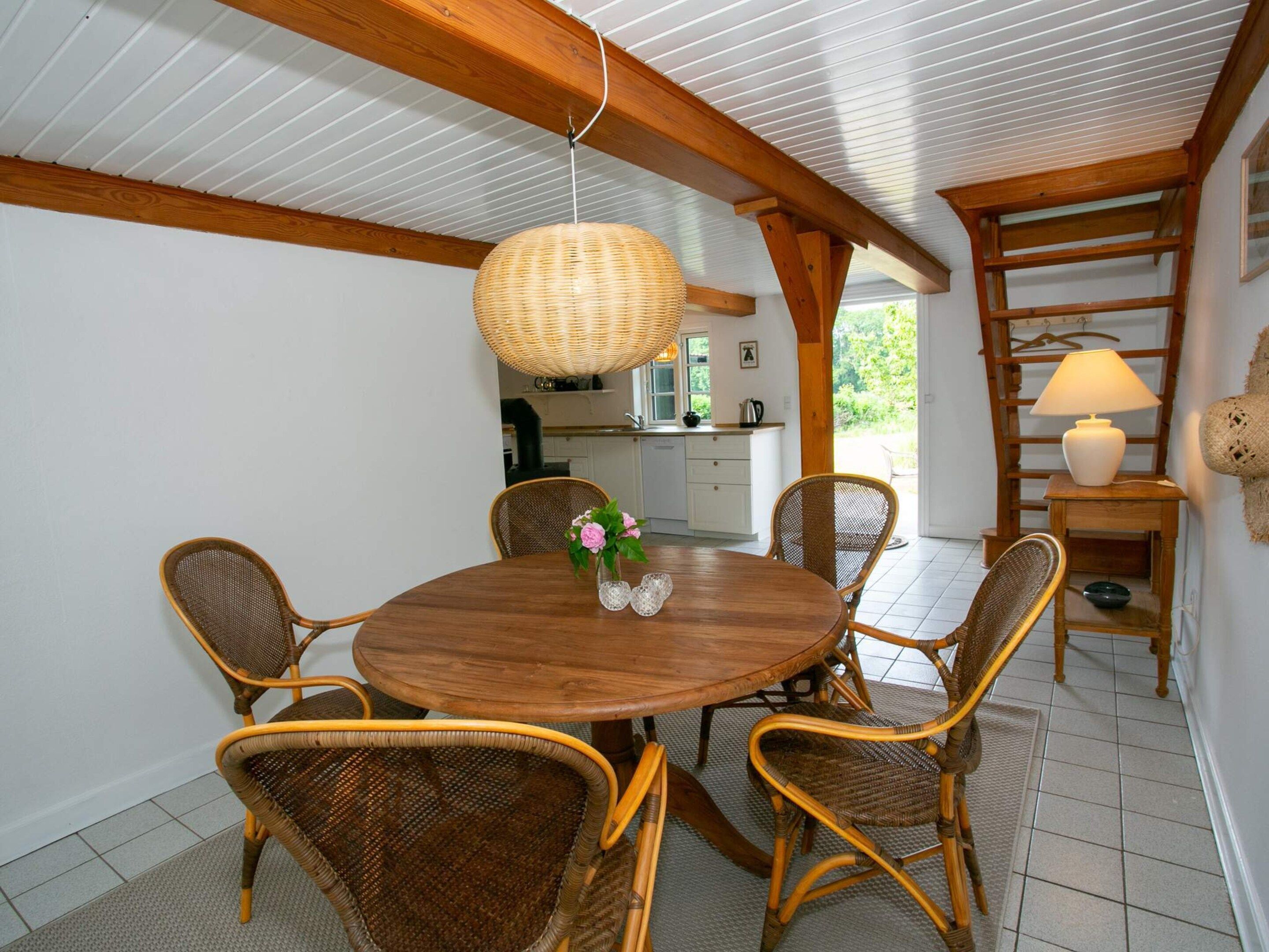 Zona pranzo con tavolo rotondo in legno circondato da sedie in vimini, lampada a sospensione in vimini e cucina sullo sfondo.