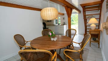Un coin repas avec une table ronde en bois entourée de chaises en osier, une lampe suspendue en osier et une cuisine en arrière-plan.
