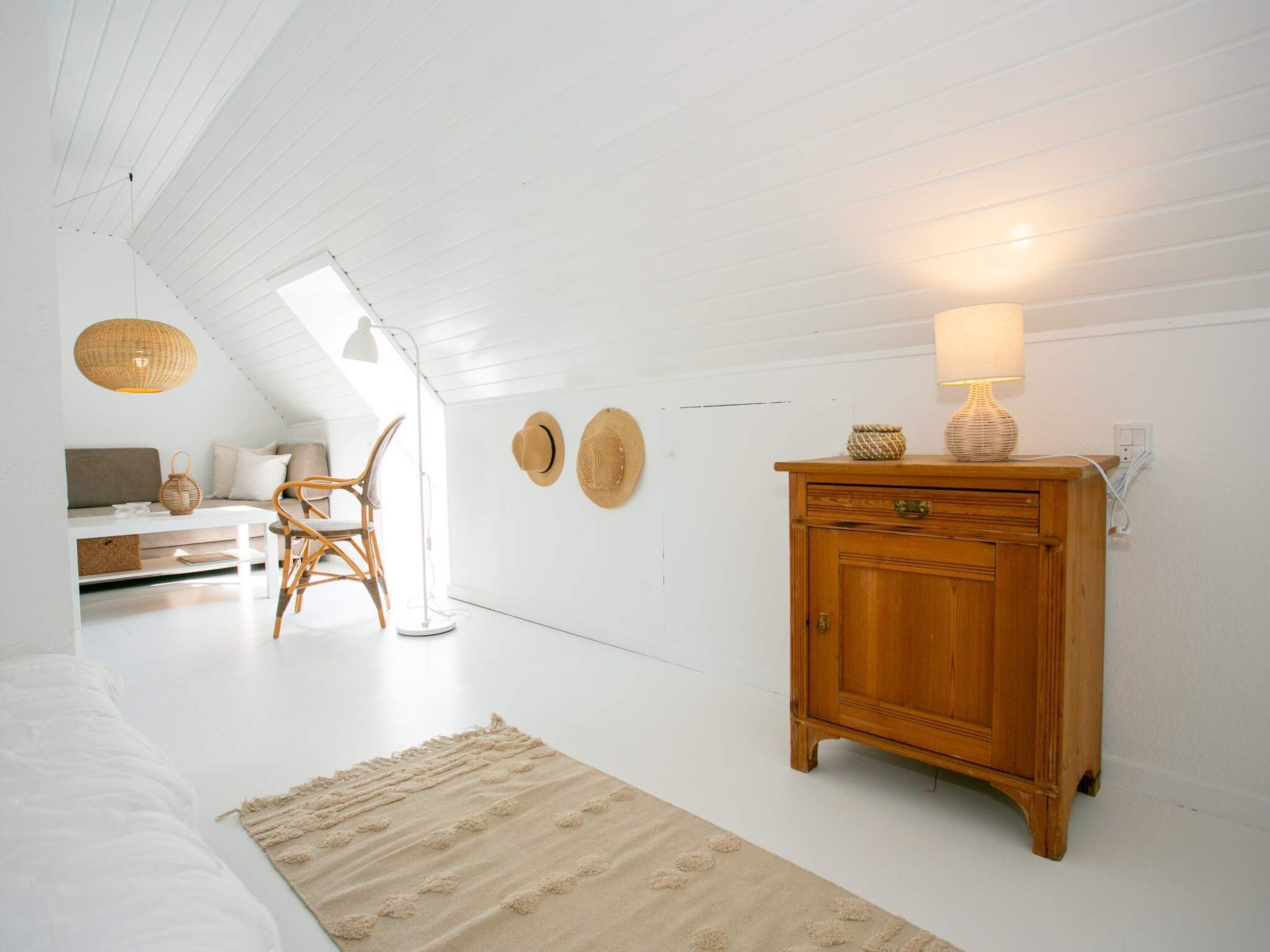 Une pièce avec un plafond en pente, une armoire en bois, une lampe en osier, un tapis tissé et un présentoir à chapeaux fixé au mur.