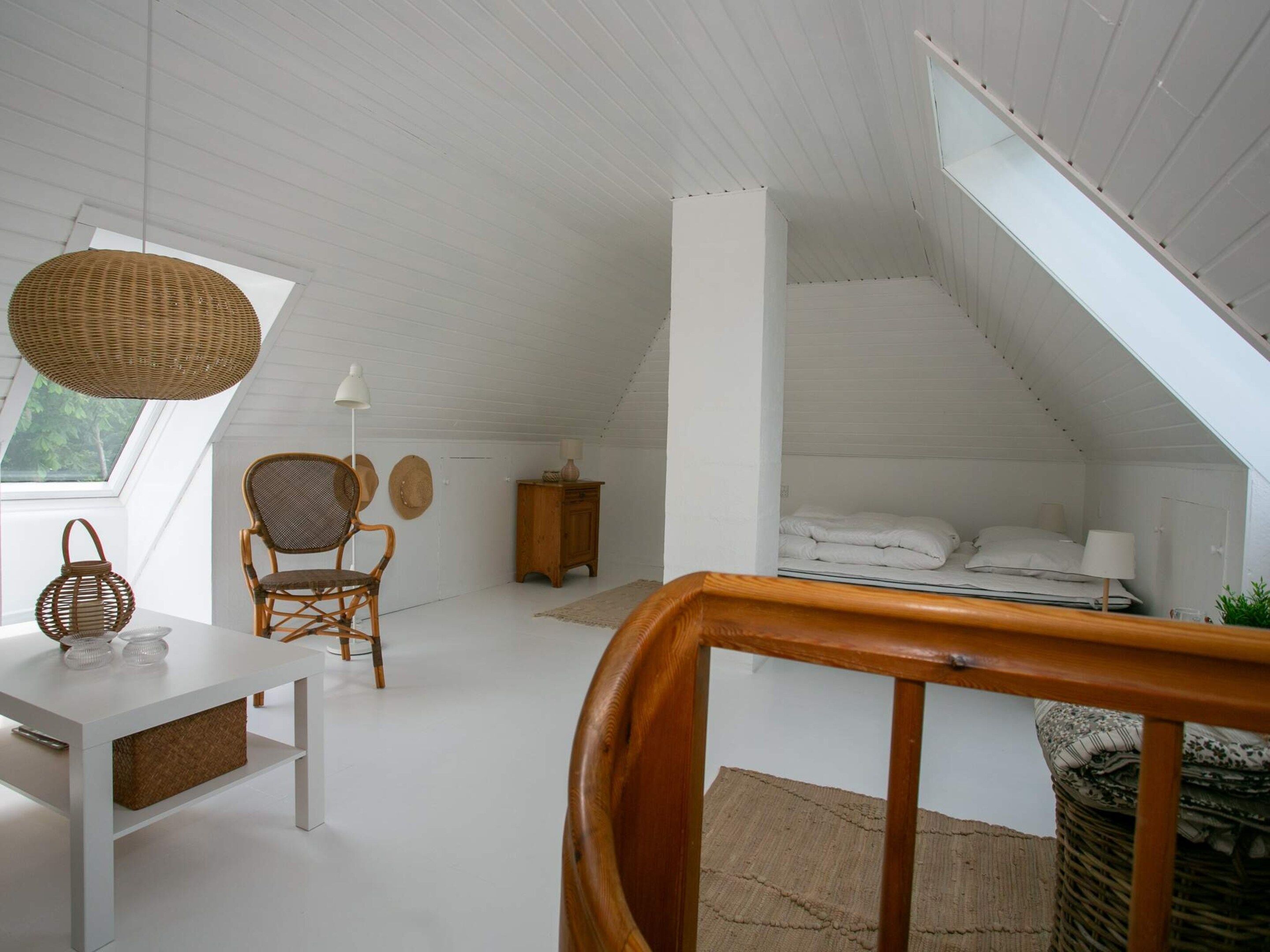 Ein Schlafzimmer im Dachgeschoss mit hohen Decken, einem geneigten Dach, einem Holzstuhl, einem kleinen Tisch und einem Bett mit weißer Bettdecke.