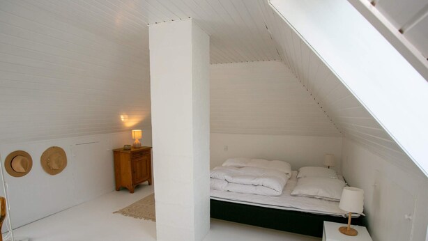 A bedroom with a sloped ceiling, a bed with white bedding, a nightstand with a lamp, and two hats on the wall. - 4 Person Holiday Home in Rudkobing (Rudkobing)