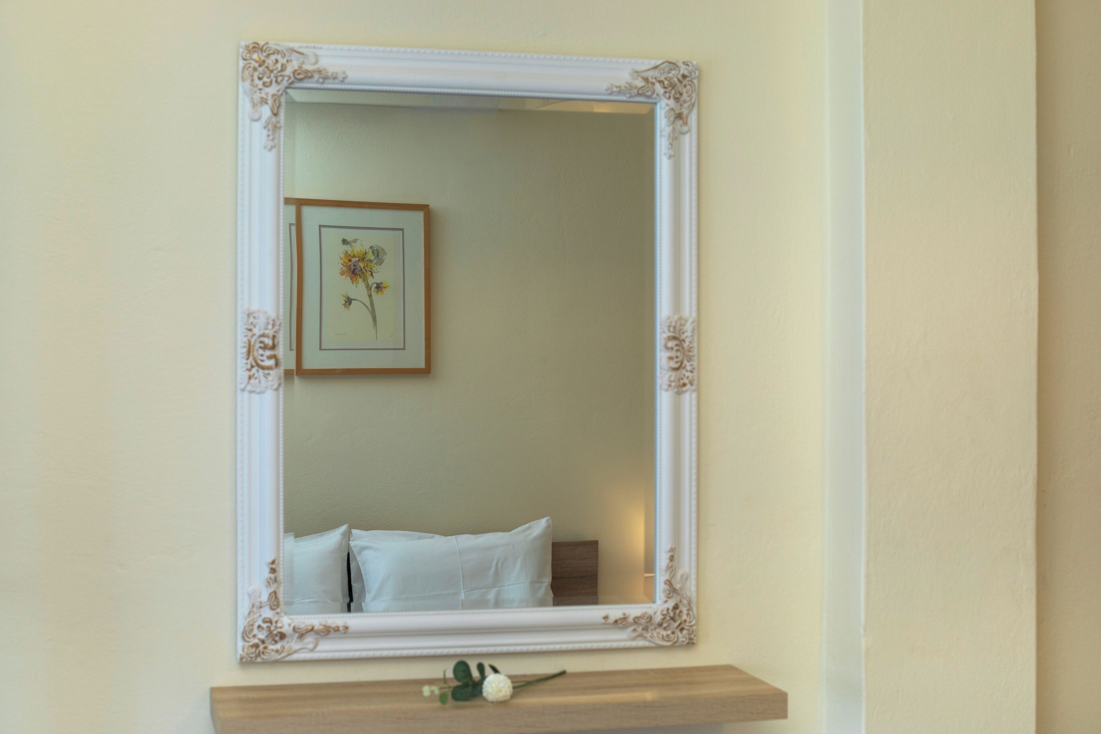 A framed mirror with a decorative border, a small framed picture of flowers, and a white pillow on a wooden shelf below.