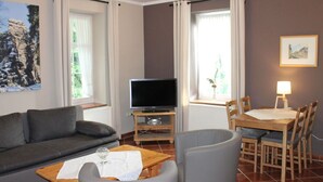A living room with a sofa, a coffee table, a TV, and a dining area with chairs and a table. - Holiday Apartment Comfortable Vacation Home (Annaberg-Buchholz)