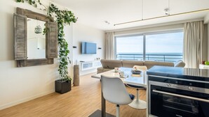 A modern living room with a sofa, a dining table with chairs, a TV, and a large window with a view of the sea. - Apartment by Zeedijk With Sea View (Koksijde)